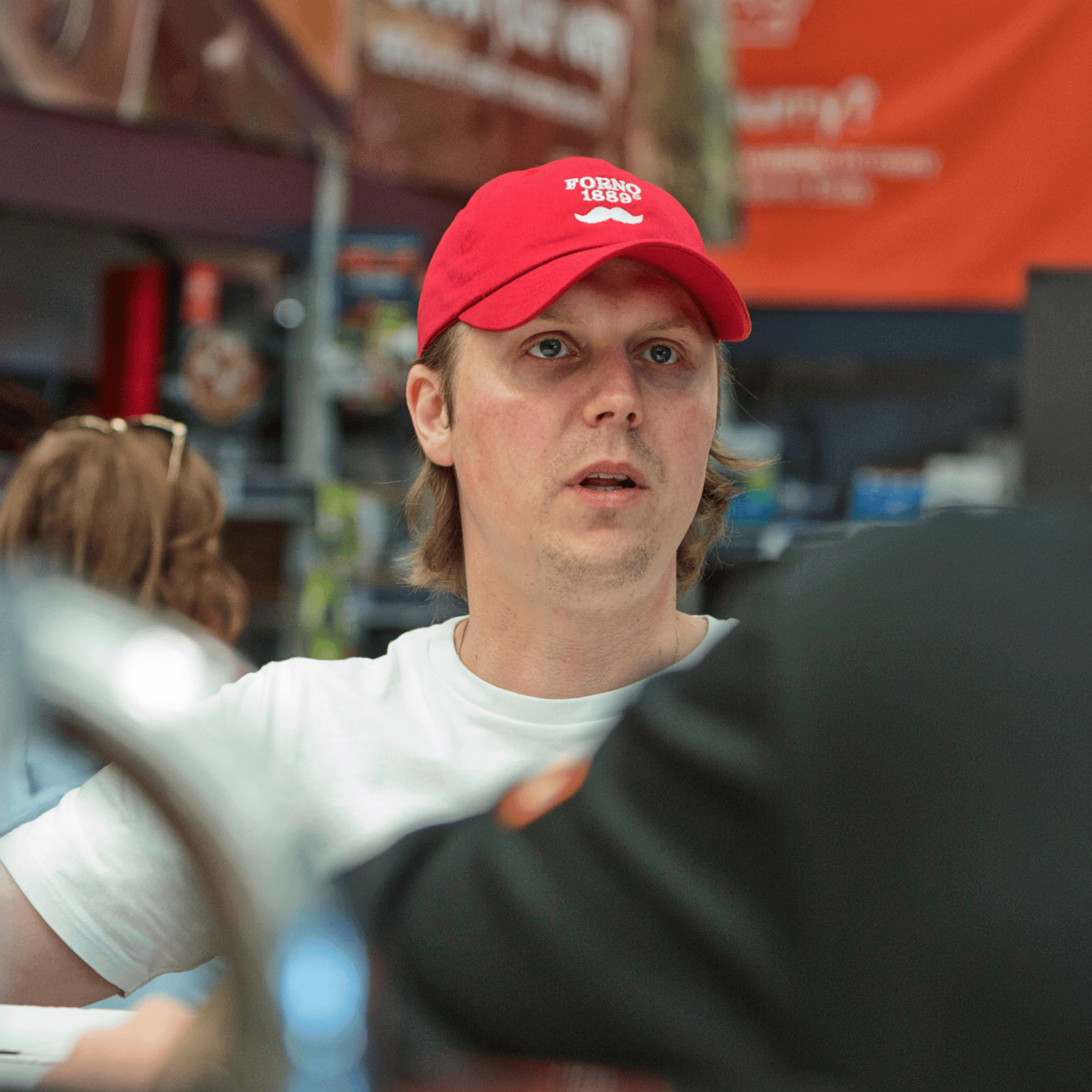 Janis Vitols wearing a red cap with a Forno1889 logo, sitting outdoors with blurred background